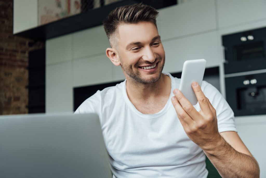 Selective focus of smiling freelancer holding smartphone near laptop at home